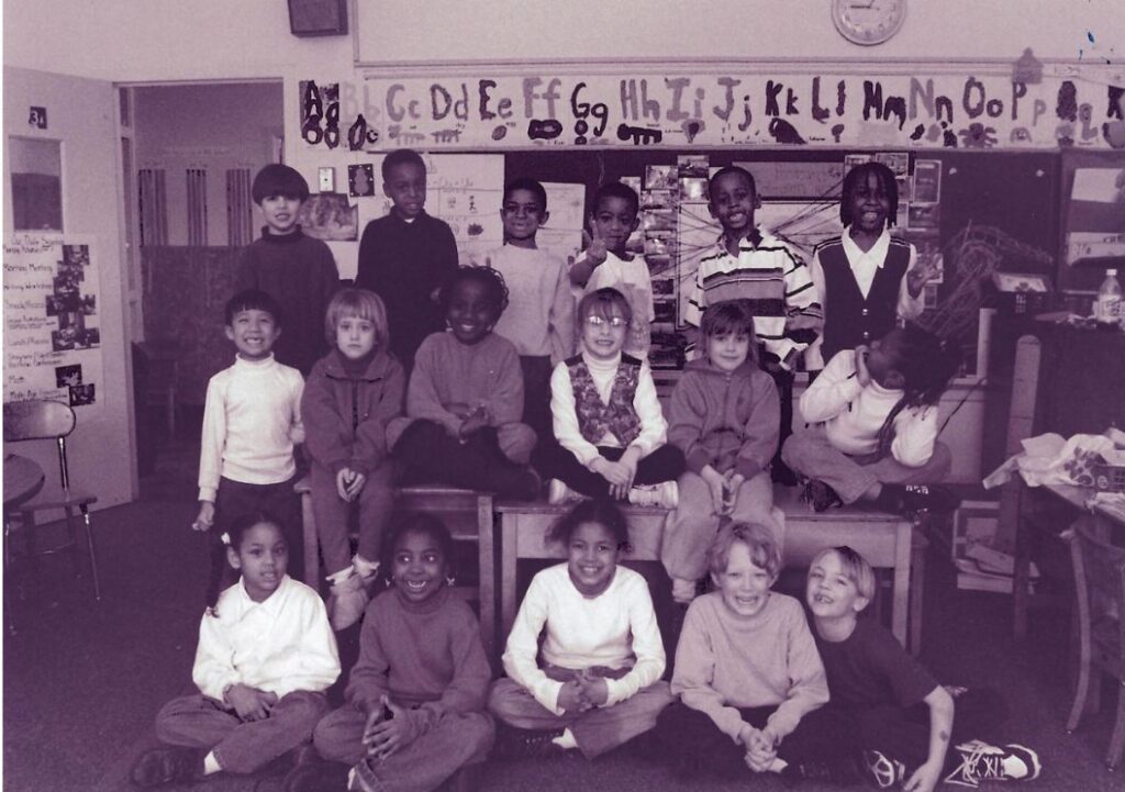 A black and white photograph of the first-ever first-grade class at Neighborhood House Charter School in Boston, Massachusetts. The classroom walls are decorated with an alphabet chart and student artwork. The diverse group of students, some seated on benches and others on the floor, are smiling at the camera.