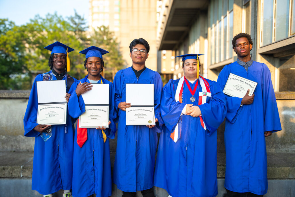 Five male graduates in blue caps and gowns holding diplomas at the Match Charter Public High School commencement.