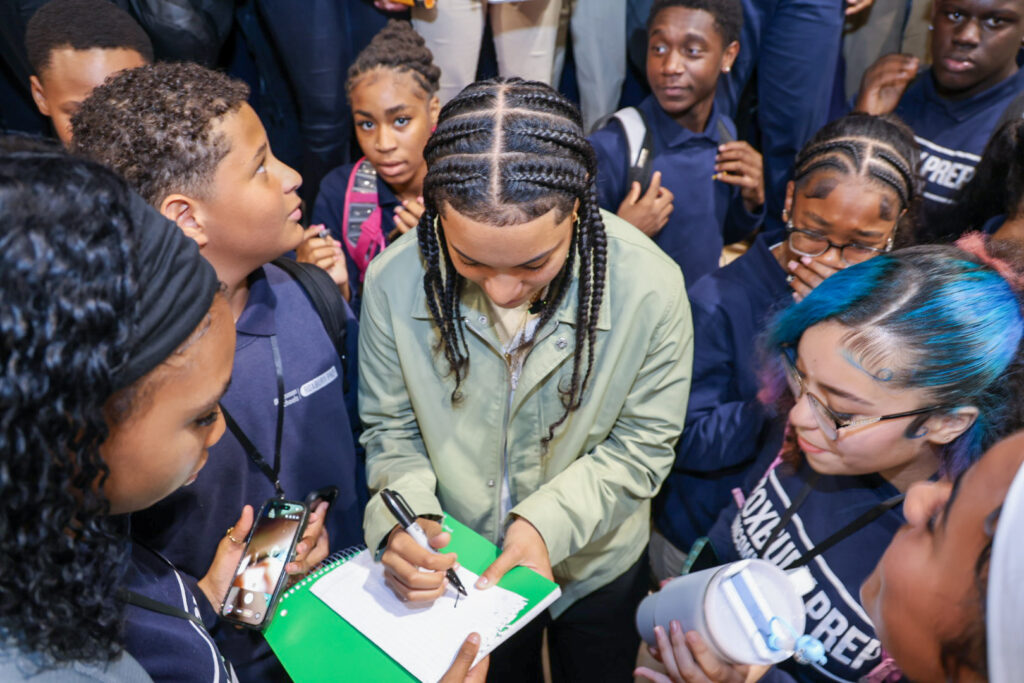 Seattle Storm guard and Roxbury Prep alumna Taina Mair signs an autograph for a student in a green notebook while surrounded by a crowd of Roxbury Prep students in navy uniforms during her homecoming celebration.