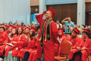 A Libertas Academy Charter School graduate in a red cap and gown, wearing a Massachusetts Army National Guard stole, salutes the crowd as classmates applaud at commencement.